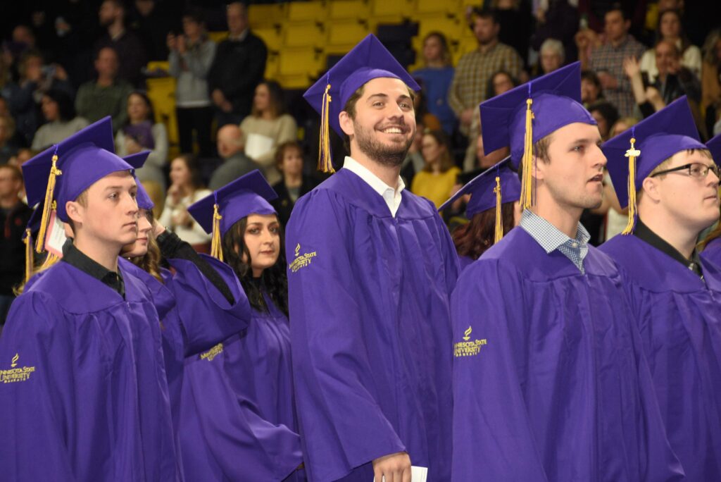 Line-up of Minnesota State Mankato graduates in purple commencement regalia. One taller student looking off in the distance with a huge smile on his face.