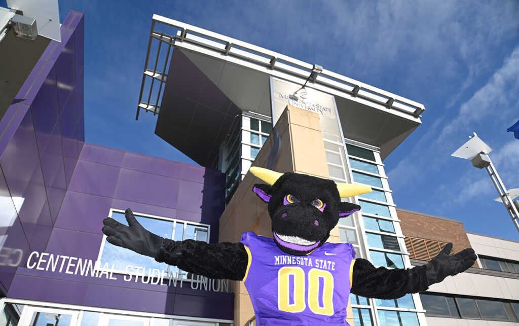 Stomper is the bull mascot for the Minnesota State Mankato Mavericks. In this photo, Stomper stands in front of the Centennial Student Union with arms outstretched wearing a Minnesota State jersey with jersey number double zero 00.